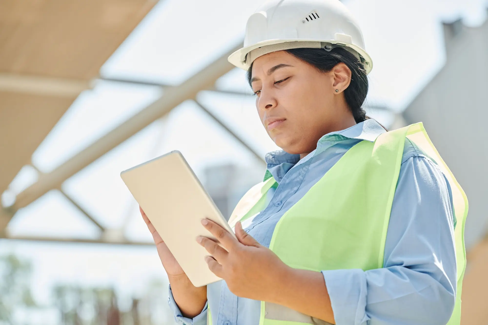 A site supervisor in a hard hat and hi-vis vest reviewing a tablet on a construction site.